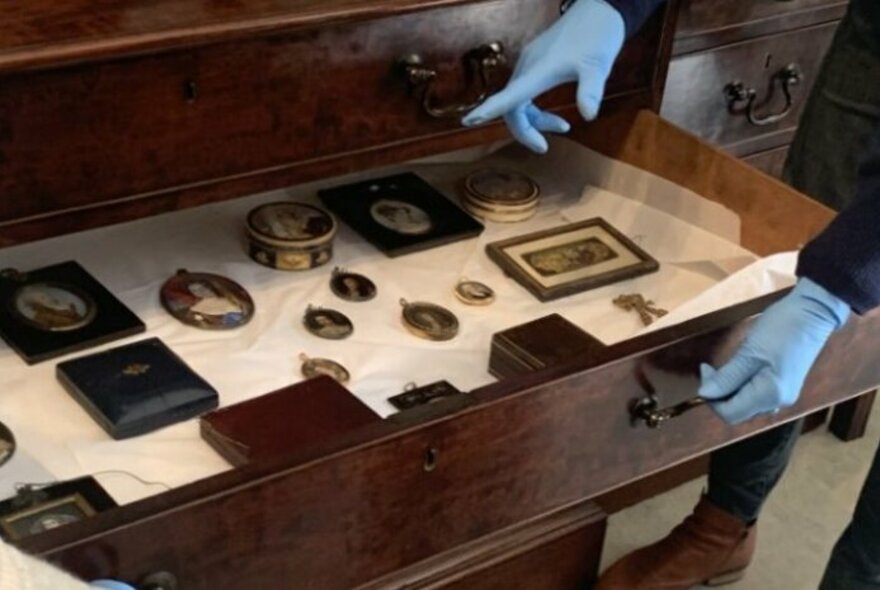 A wooden display case containing a number of small items, being opened by hands wearing blue rubber gloves.