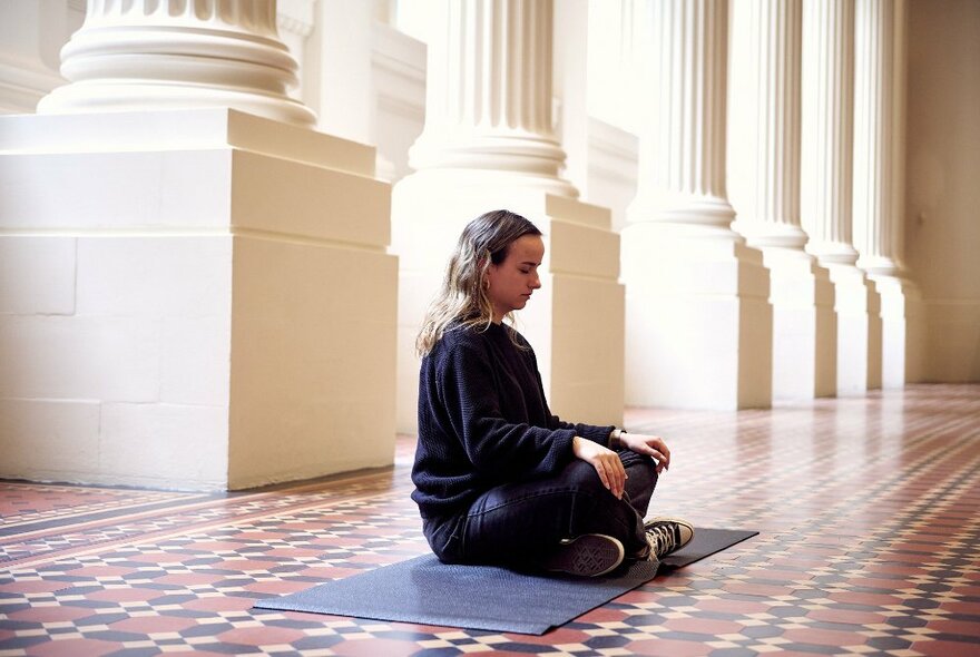A woman sitting meditating on a tessellated tile floor with white columns behind her.