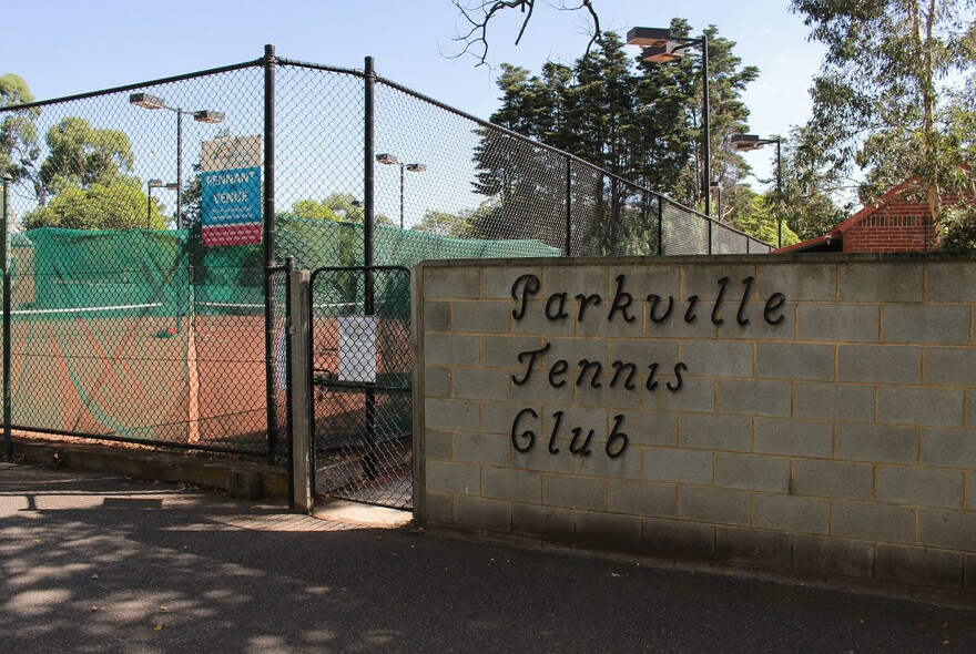 Exterior of Parkville Tennis Club showing wall, signage, gate, a fenced tennis court and trees in the background.
