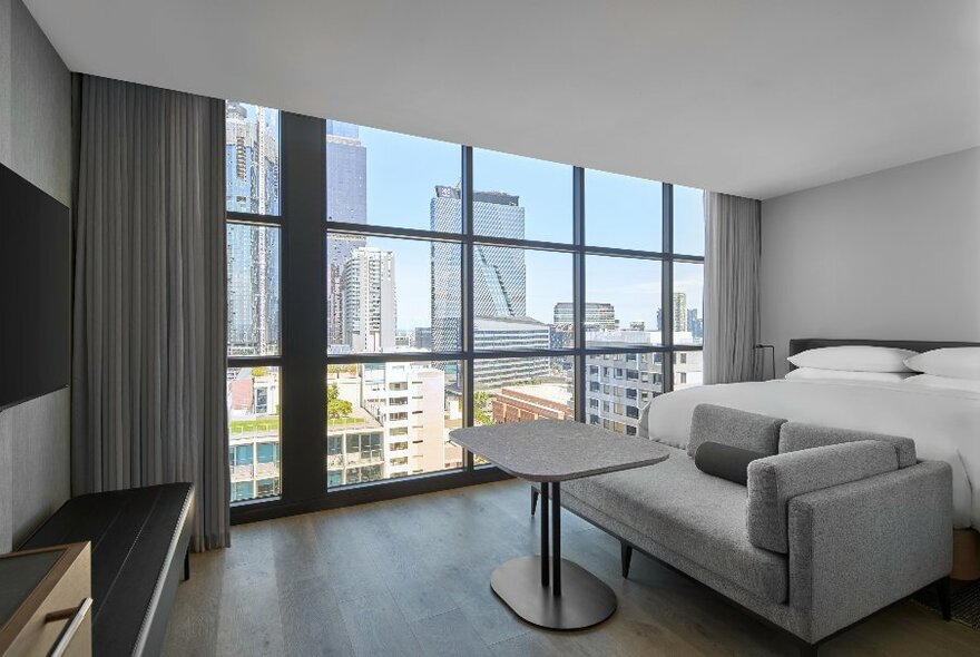 A deluxe room at the Courtyard Melbourne hotel with king size bed, grey couch and wide windows overlooking the city skyline.