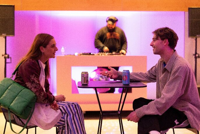 Two people sit opposite each other at table with two cans of drink. A DJ is in the background with a pink light cast on the room. 