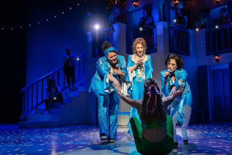 Four women on a theatre stage, all wearing blue costumes, three of them singing into microphones they hold in their hands.