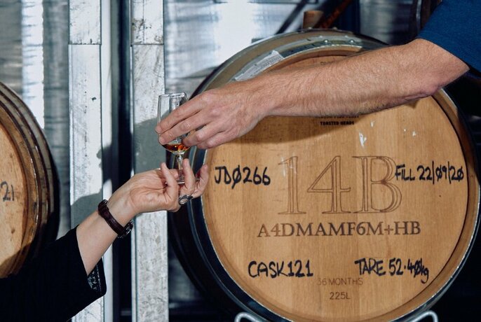 An arm reaching out to place a glass into another person's waiting hand, in front of an oak wine barrel.