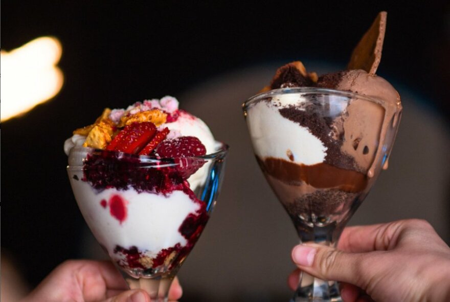 Two hands holding ice-cream desserts, one chocolate and the other with berries.