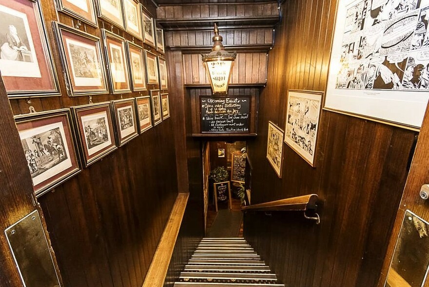 Dark wood-panelled stairs leading down to a bar, walls lined with framed artworks, lit by heritage lamp.