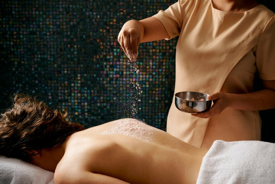 A therapist sprinkling a beauty product on the bare back of a customer lying face-down on a table, their lower torso wrapped in a white towel.