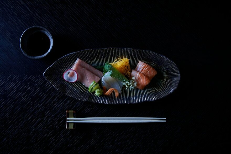 Oval plate with an array of raw fish and sashimi, pickled vegetables and chopsticks on a dark table.