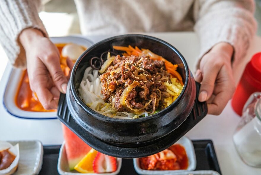 Hands holding a clay pot filled with Korean beef.