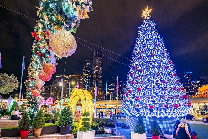 Night view of Christmas decorations at Fed Square including a large illuminated tree and giant hanging baubles, with city buildings in the background.