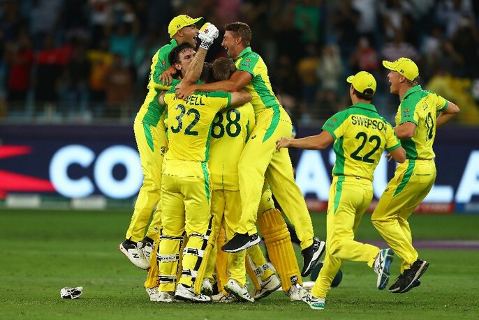 The Australian T20 men's cricket team wearing their distinctive yellow and green uniforms, in a group hug on the cricket field, spectators visible in the background.