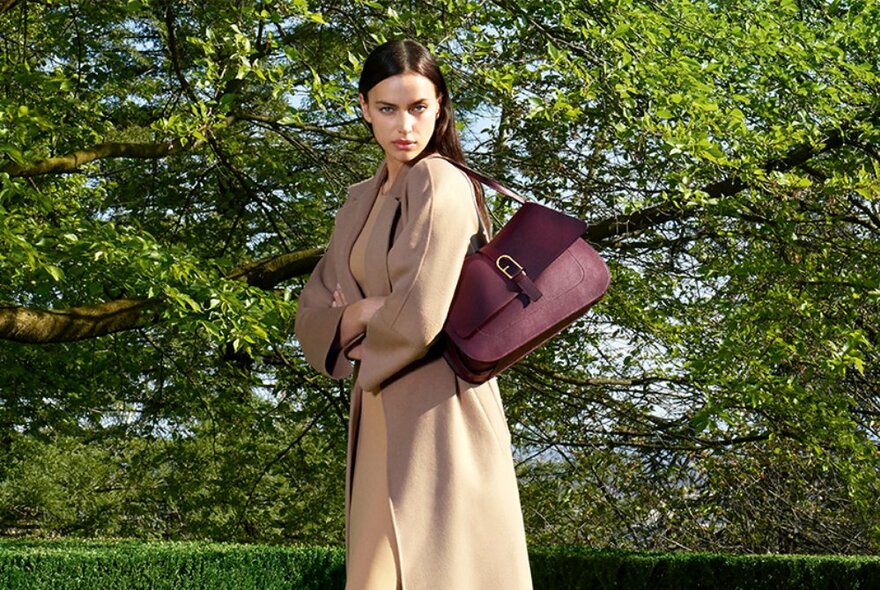 Model in a camel coloured coat, carrying a burgundy handbag over her shoulder, standing outside in a park setting.