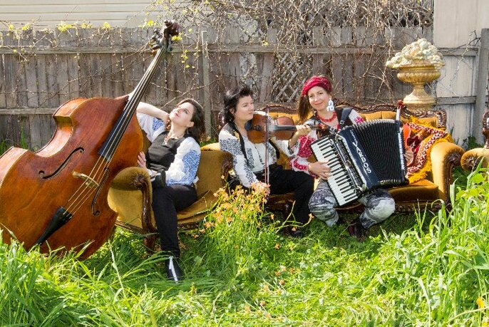 Three musicians of the musical group Vardos sitting on grass outdoors, holding their instruments of a piano accordion, a violin and a double bass.