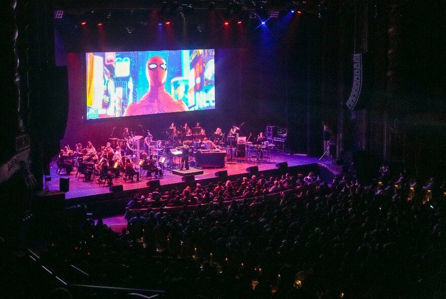 Looking down on a stage with a full orchestra in front of a large movie screen showing Spiderman. 