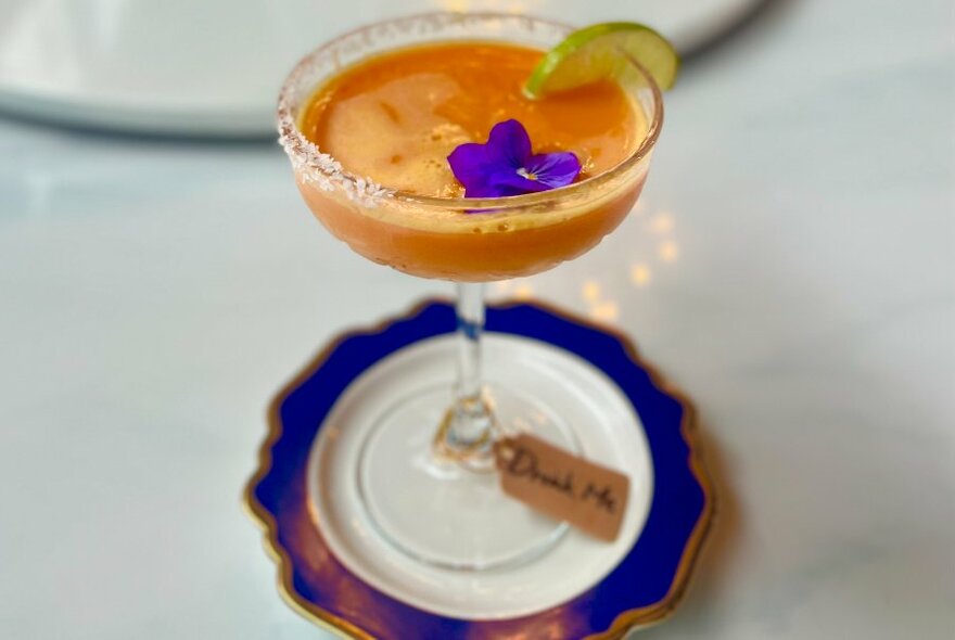 An orange cocktail with a purple violet and slice of lime on a purple saucer.