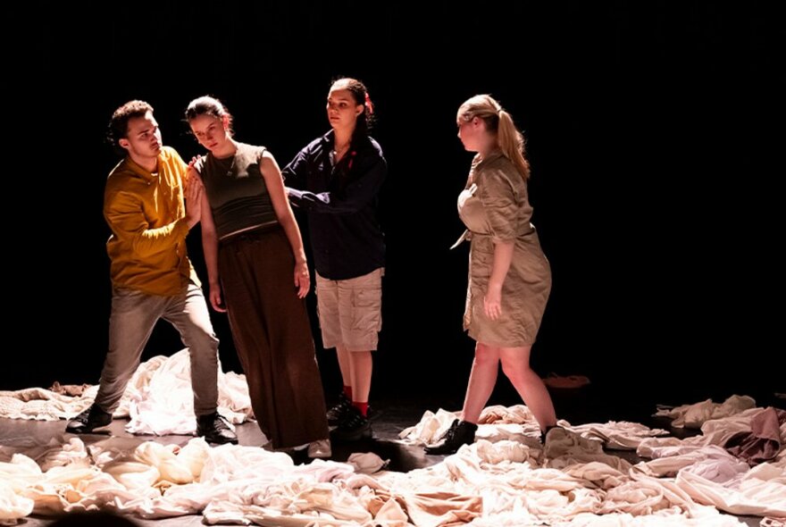 Four performers, one leaning and being held by others, on stage under a light with material strewn over the floor. 