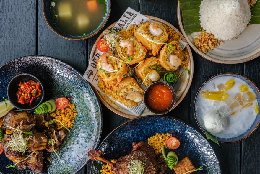 An overhead view of plates of Indonesian food, including  meats, rice, soup and a shaved ice milky dessert.