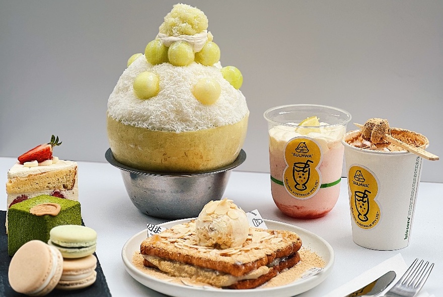 Food arranged on a table that includes shaved ice dessert topped with melon balls, mini cakes, toast, beverages, and macarons.