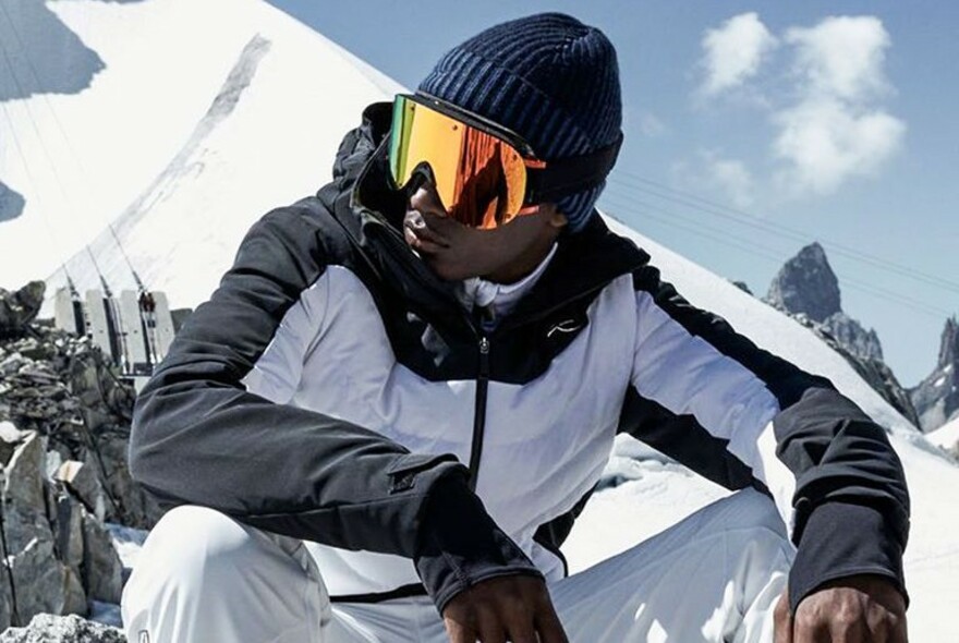 Man wearing a black and white ski jacket, ski goggles and a beanie, with a snow covered mountain in the background.