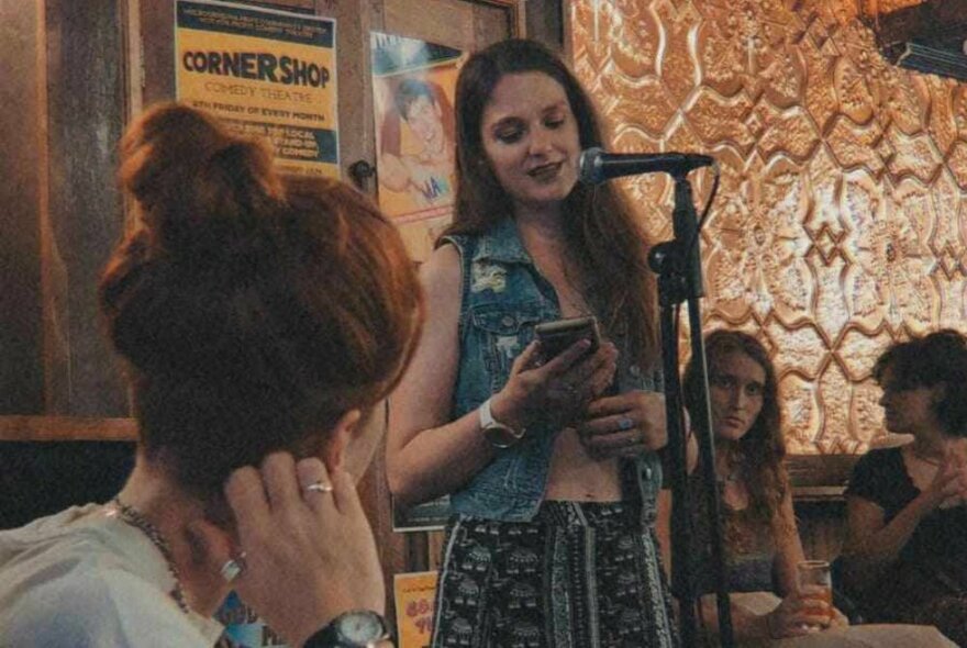 A young woman standing up in a small space and reading from her phone into a microphone while others look on. 