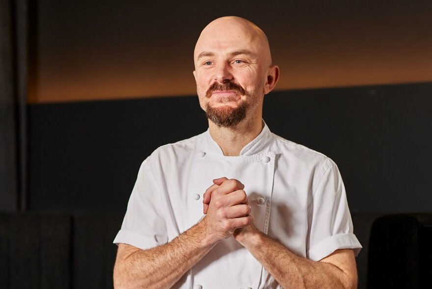 Man wearing chef's clothing with a bald head and goatee, clasping his hands together, smiling.