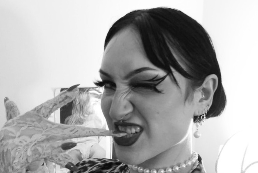 Black and white image of a woman biting her nails through lacy gloves, winking, with winged eye make-up.