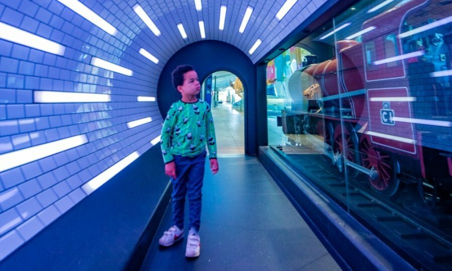 A child walking through a blue-lit tunnel looking at a train. 