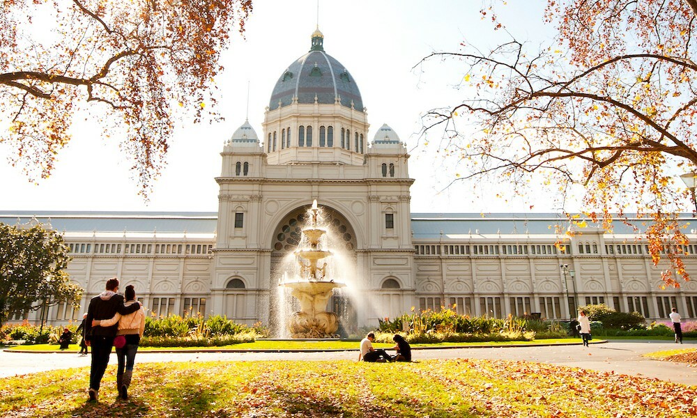 A couple standing in front of the Royal Exhibition Building, surrounded by trees.
