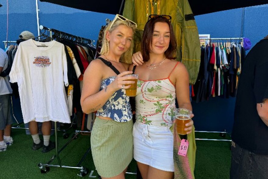 Two people posing with drinks in front of racks of secondhand clothing.