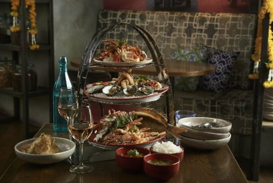 Three tiers of a seafood platter with side bowls of rice, dipping sauce and two glasses of white wine on a table.