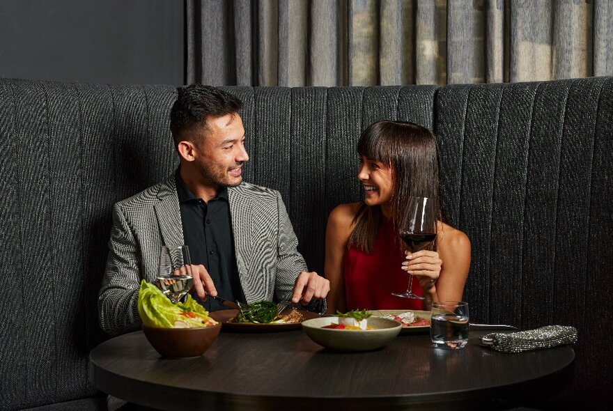 A couple seated at a restaurant banquette, eating an array of dishes and drinking red wine.