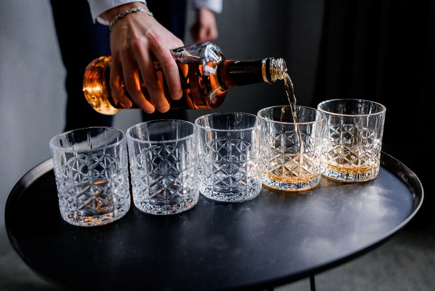 Hand pouring whisky into five cut-crystal tumbers lined up on a dark tray table.