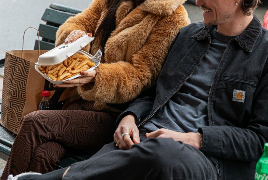 Two people seated on bench, seen from  chin down, one on left opening container of hot chips.