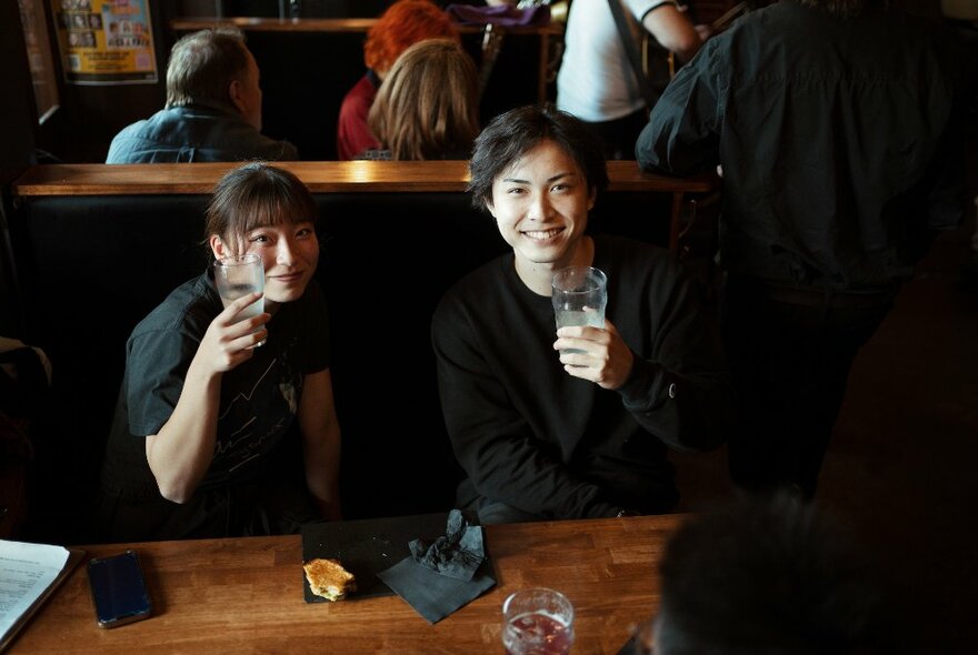 Two people smile while seated at a wooden table and holding up a drink. 