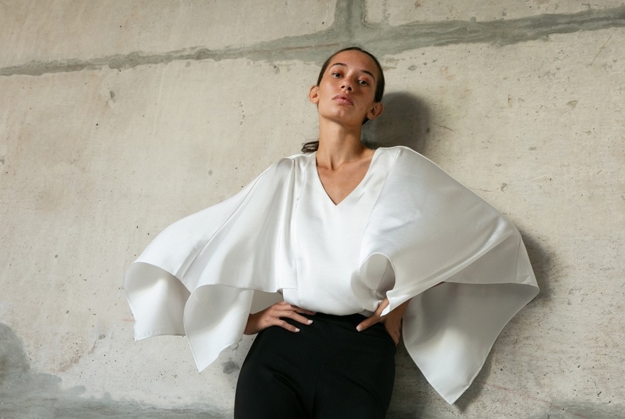 Female model wearing a white top with large billowing open sleeves, tucked into black trousers, posed against a concrete wall.