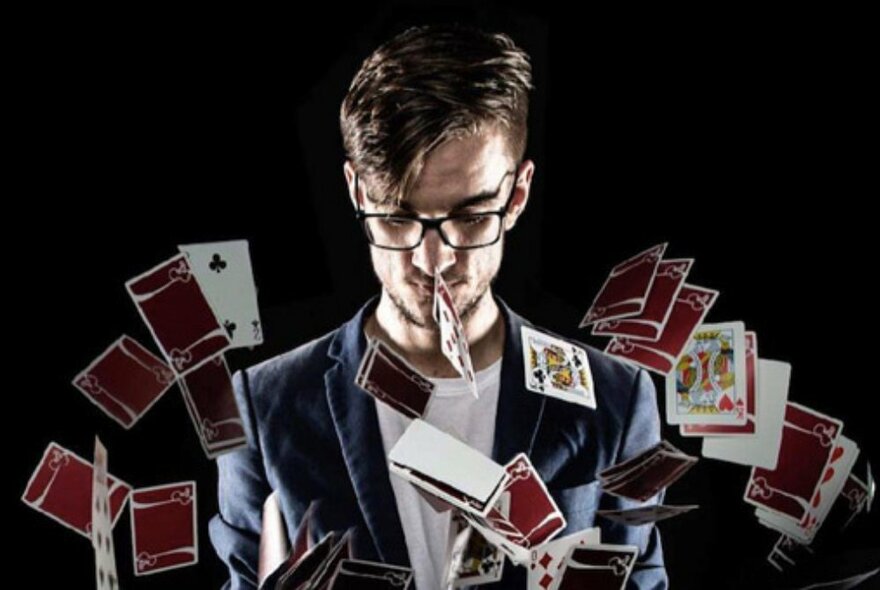 Magician with beard and glasses, looking down as playing cards fill the air around him.