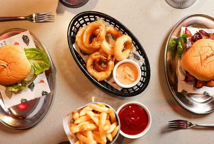 Onion rings, fries and two different types of burgers on a light-coloured table, from above.