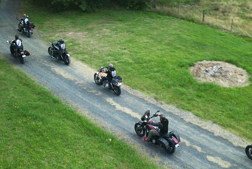 Five motorcyles driving along a small gravel road flanked by green grass.