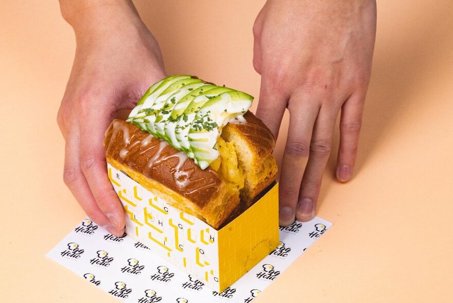 A pair of hands resting on a small yellow takeaway box, which contains an oversized toasted sandwich with a scrambled egg filling.