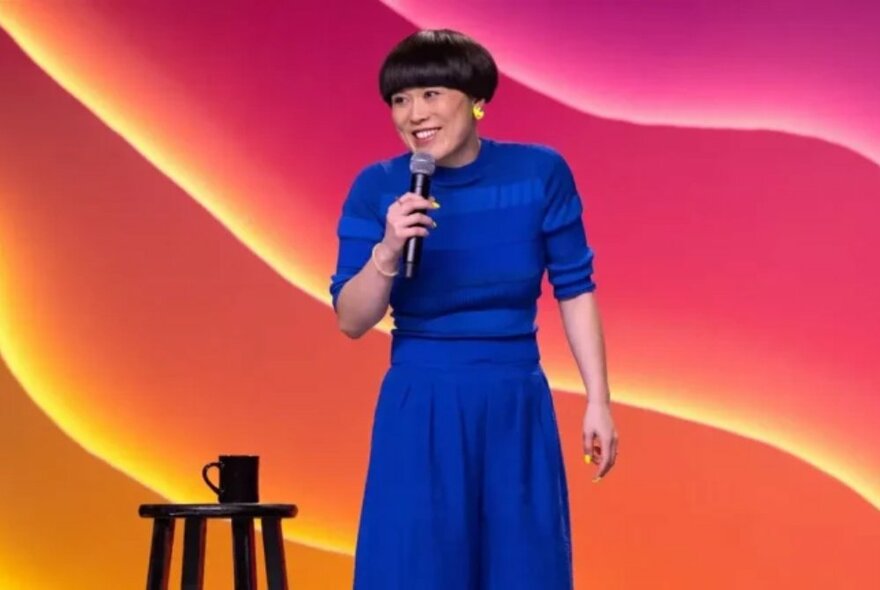 Comedian Atsuko Okatsuka with a bowl haircut, holding a microphone and performing stand-up comedy on a stage.