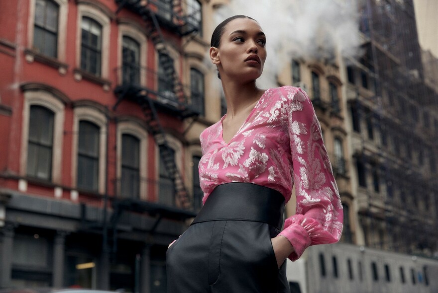 Model in bright pink blouse and high-waisted black pants, with cityscape behind.