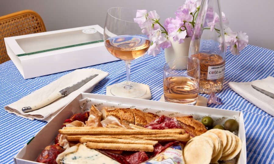 A charcuterie and cheese platter in a box next to a glass of rose.