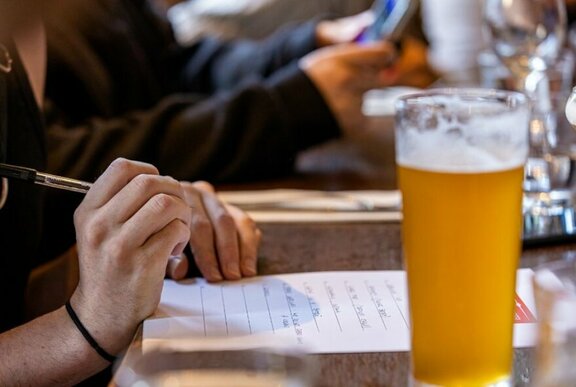 A hand holding a pen and writing answers on a sheet of paper, with a pint of beer on the table.