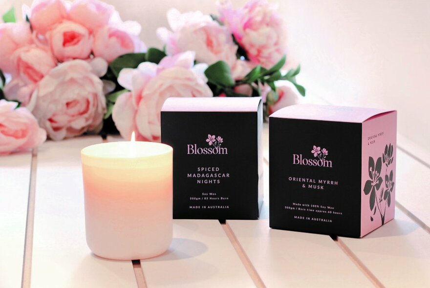 A lit candle beside two gift boxes with pink roses in the background. 