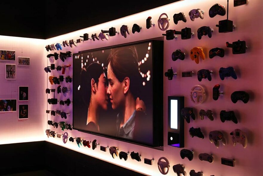 Screen showing two people embracing, surrounded by games consoles mounted on a gallery wall.