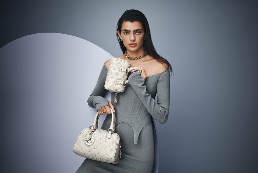 Model in grey dress holding two small, white-cream bags, against two-toned grey backdrop.