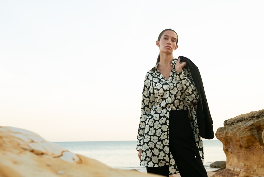 Female model wearing a floral patterned long shirt over dark trousers, posing near rocks at a beach.
