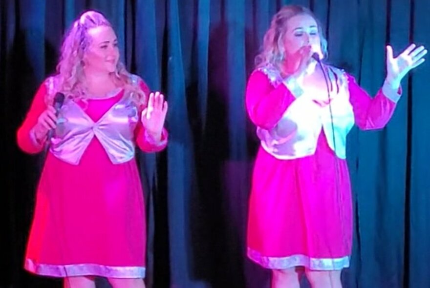 Twins performing on stage wearing bright pink dresses with silver details, singing into microphones.