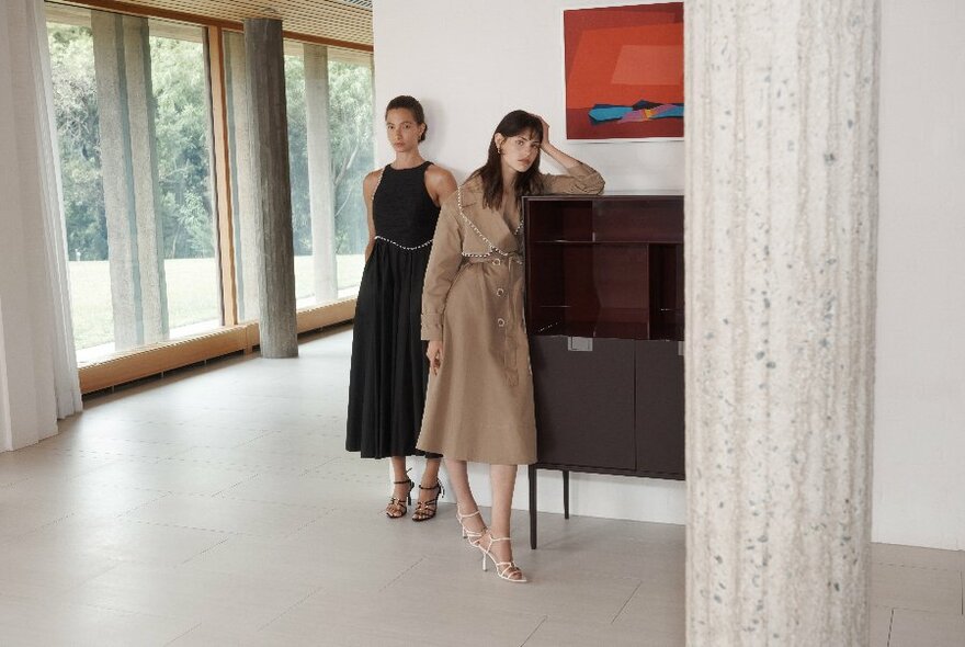 Two models, one in black, one in taupe dress, white, standing in white room, floor to ceiling windows at right, cream column to right.