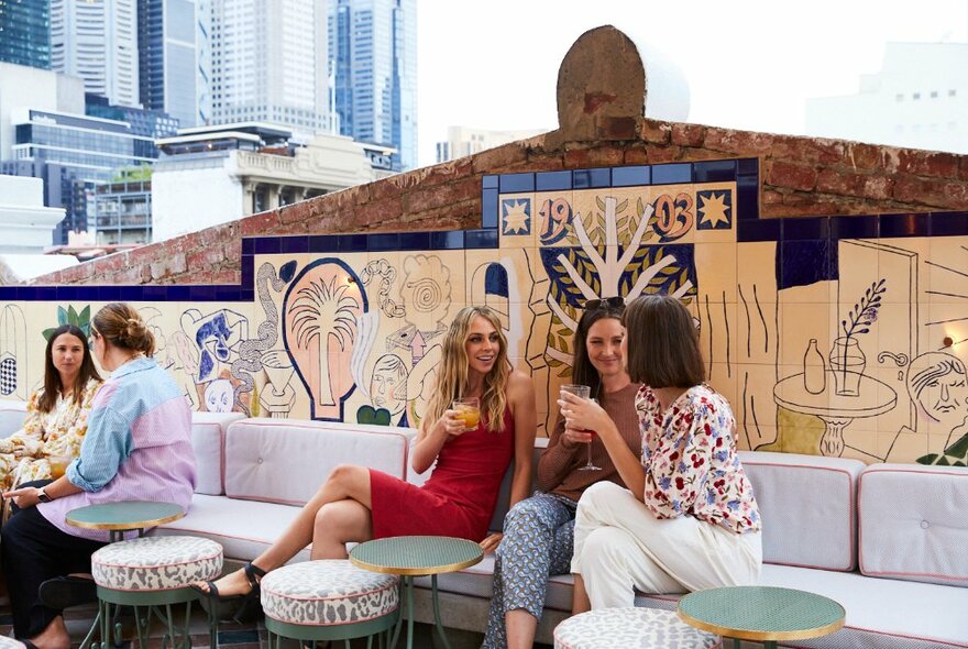 People seated on banquette couches at an open air rooftop bar with the Melbourne skyline in the background.