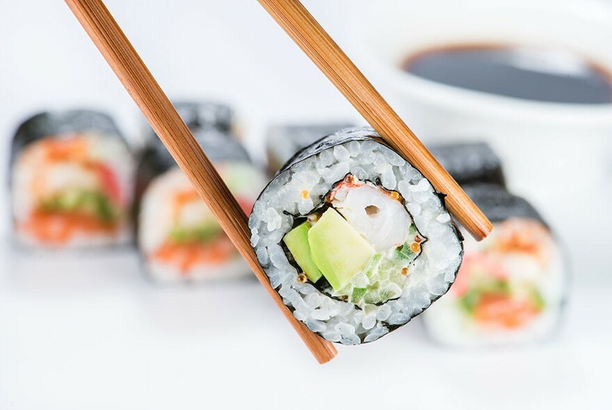 Small nori roll held up by chopsticks, with row of  nori rolls in background.